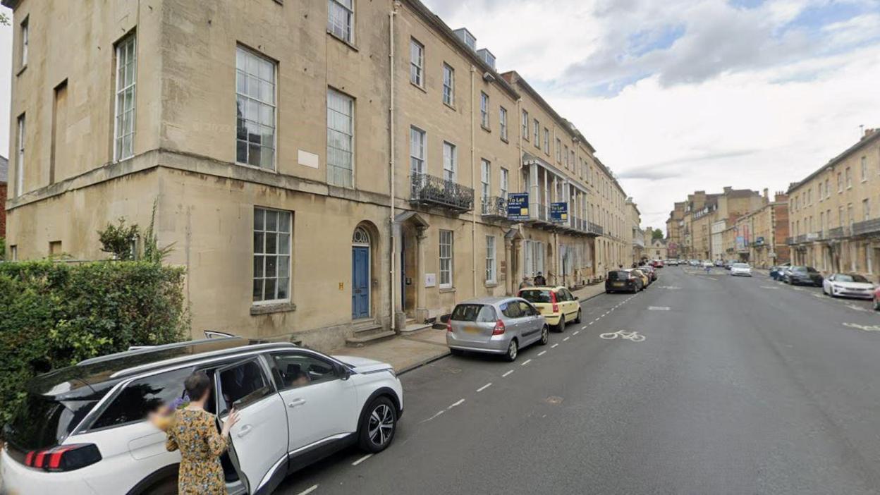 A view of Beaumont Street . There are cars parked on the sides. Someone is seen getting in to a car, holding a bouquet of flowers. It is a cloudy day.