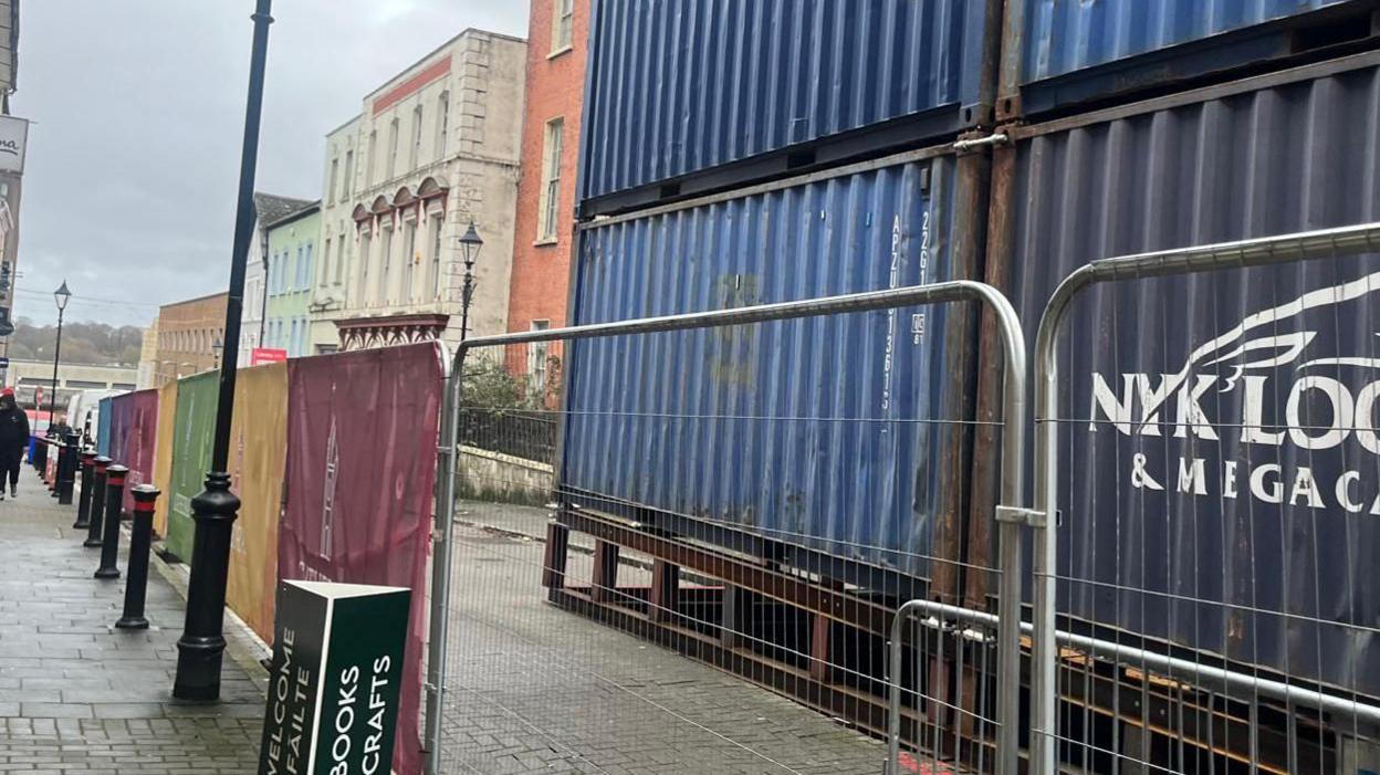 Large blue shipping containers are stacked on top of each other in front of a metal railing. A number of black lamppost are also visible along a long narrow street.