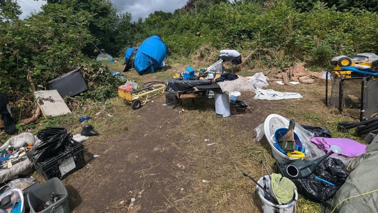 Piles of rubbish including an old watering can, a tent, and some furniture littered across a path in an overgrown park