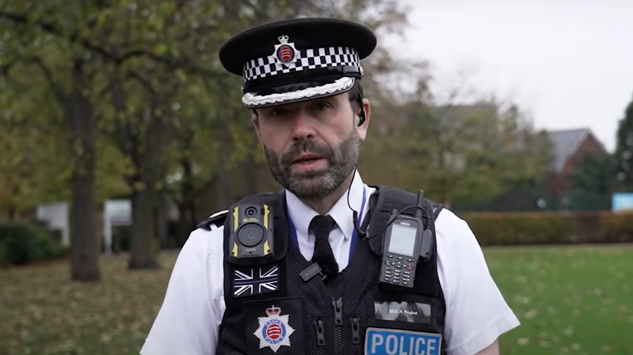 Andy Prophet in police uniform, including his black vest, body camera and radio. He is wearing his police issue hat, he has a thick beard and he is standing in a field.