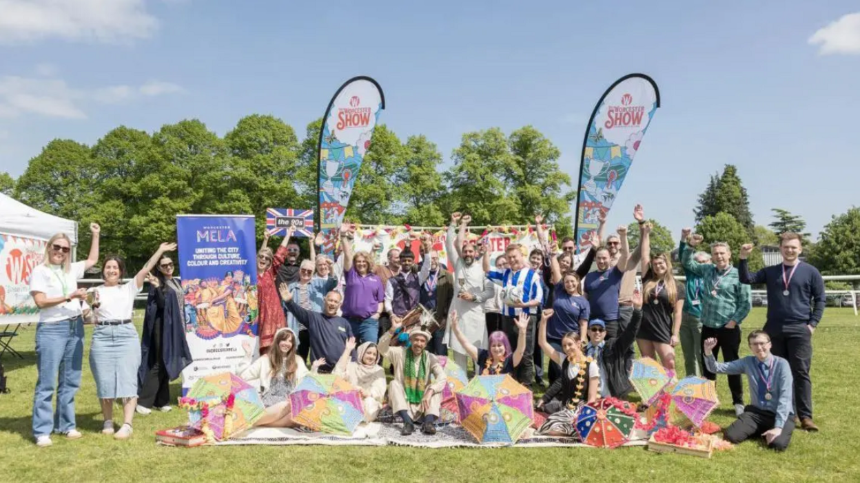 A large group of people posing for the camera on a field with flags stating Worcester Show. Several have their arms raised and most are smiling.