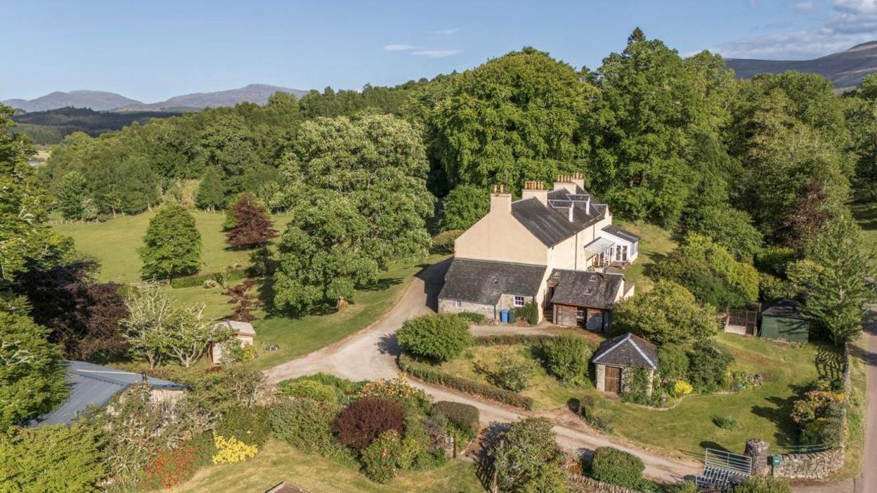 The house is of period design with a slate roof and cream-coloured walls. It is set in a Highland landscape of rolling hills. Trees surround the property.