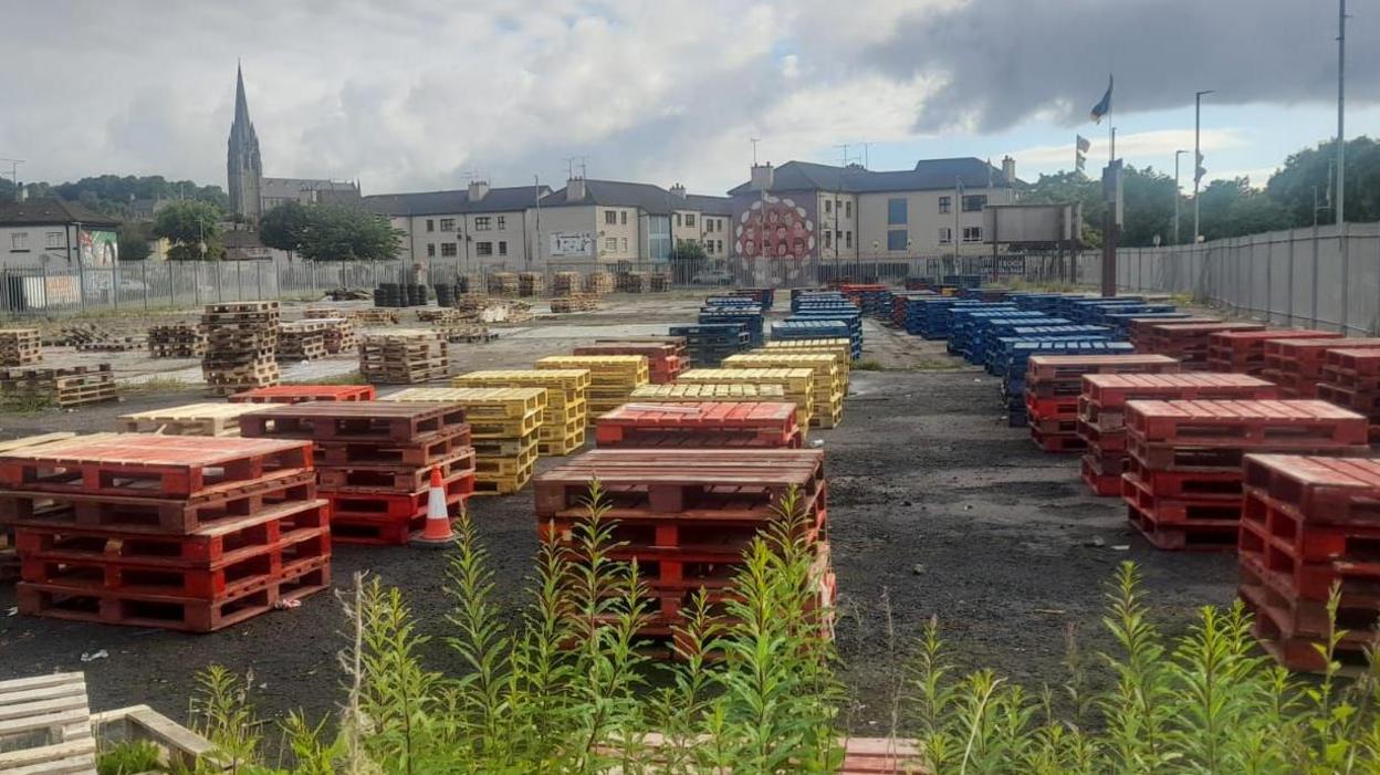 a vacant piece of land has dozens of wooden pallets stored upon it. a number of houses and a cathedral spire can be seen in the background