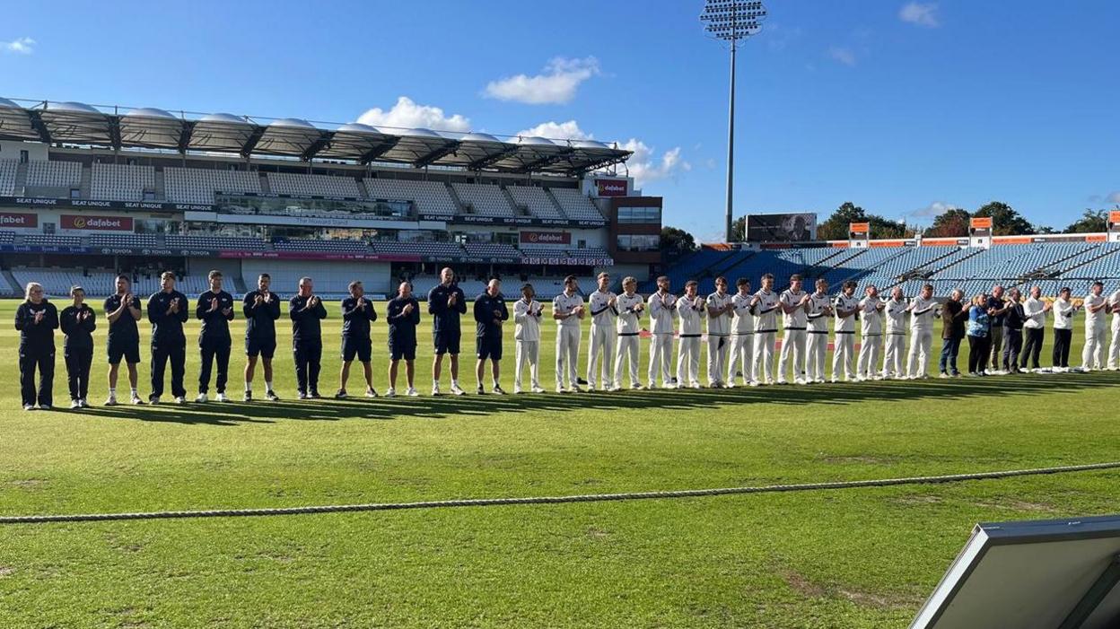 A row of around 30 people standing in a line clapping. The team, in the middle of the line, all wear white. 