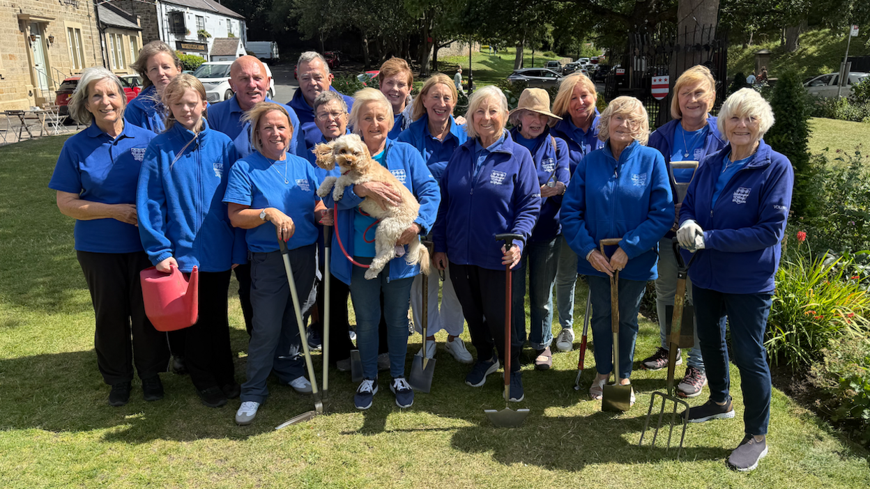 Volunteers from the Washington Village in Bloom group are wearing blue coloured shirts and fleeces and standing with various gardening equipment. One person teenager is holding a red watering can while Joan (in the centre) is holding up a blonde cavapoo dog named Bailey.
