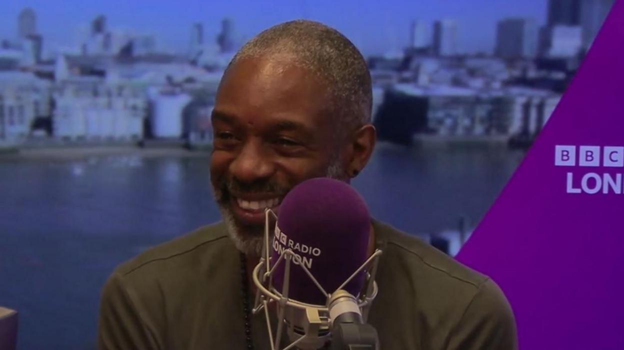 Wil Johnson in a BBC London radio studio. He wears a green t-shirt and sits behind a purple microphone.