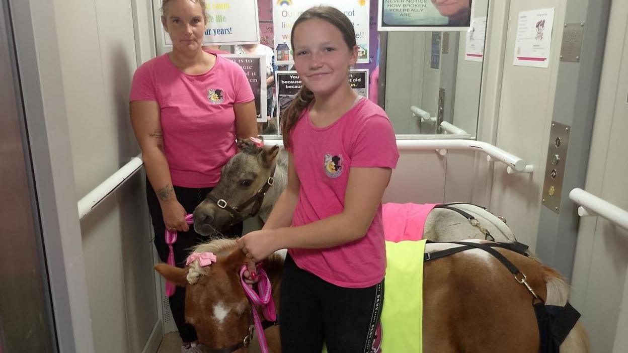 Kelly and Farrah in pink T-shirts and black leggings stand in a lift with two ponies so they can transport them down to the ground floor of the care home. 