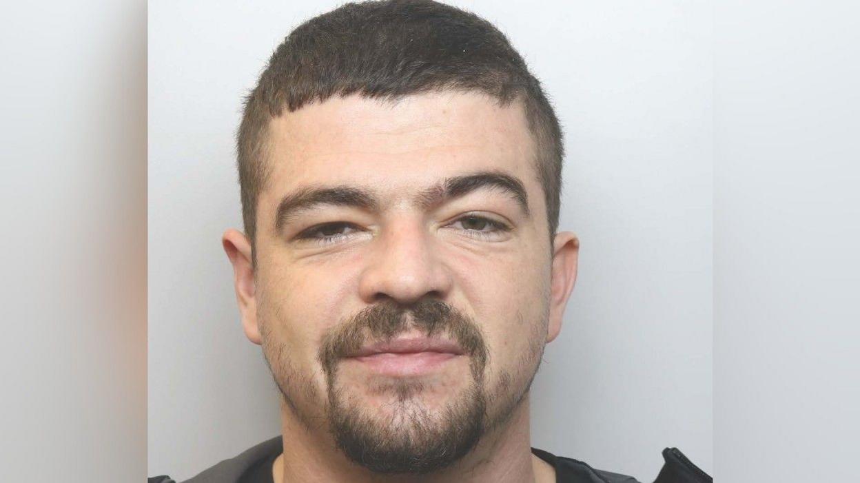 A police mugshot of a man with short dark hair and a dark goatee-style beard.