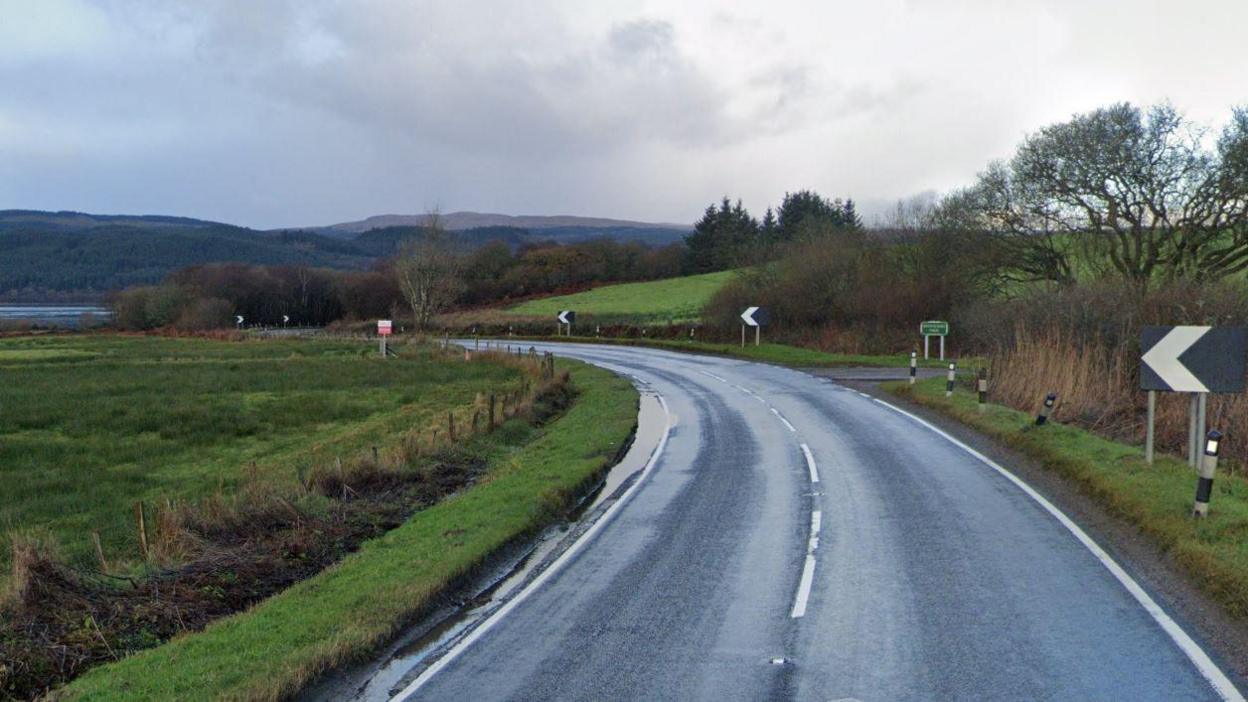 A bend in a single carriageway road surrounded by fields and some trees.