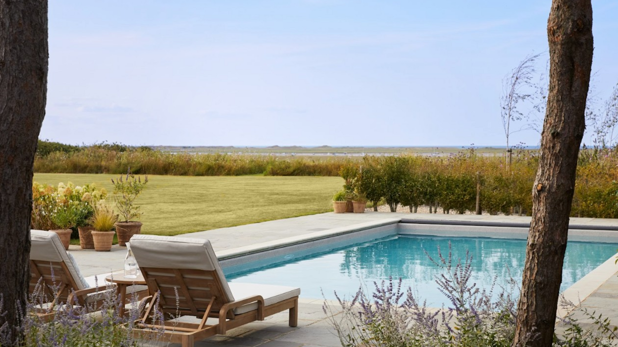 Two wooden sun loungers with beige cushions beside a swimming pool, with a lawn to one side and a view beyond of Blakeney Point 