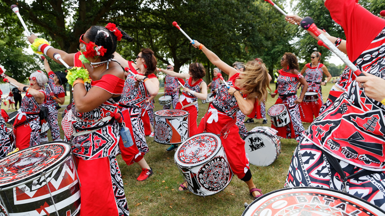 Women dressed in red, black and white costumes performing while playing drums at Leicester Caribbean Carnival