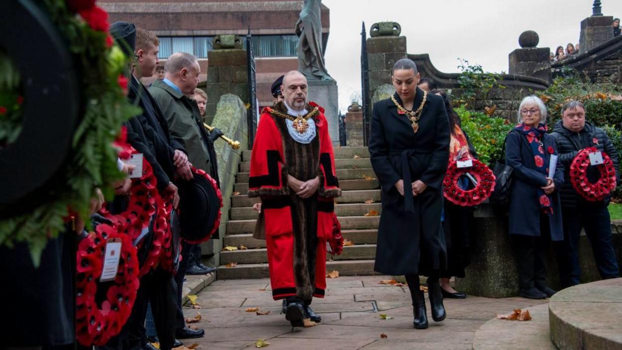 A man with a white beard and long red coat walking alongside a woman with dark hair and a dark coat with other people in dark clothing on either side holding wreaths of poppies