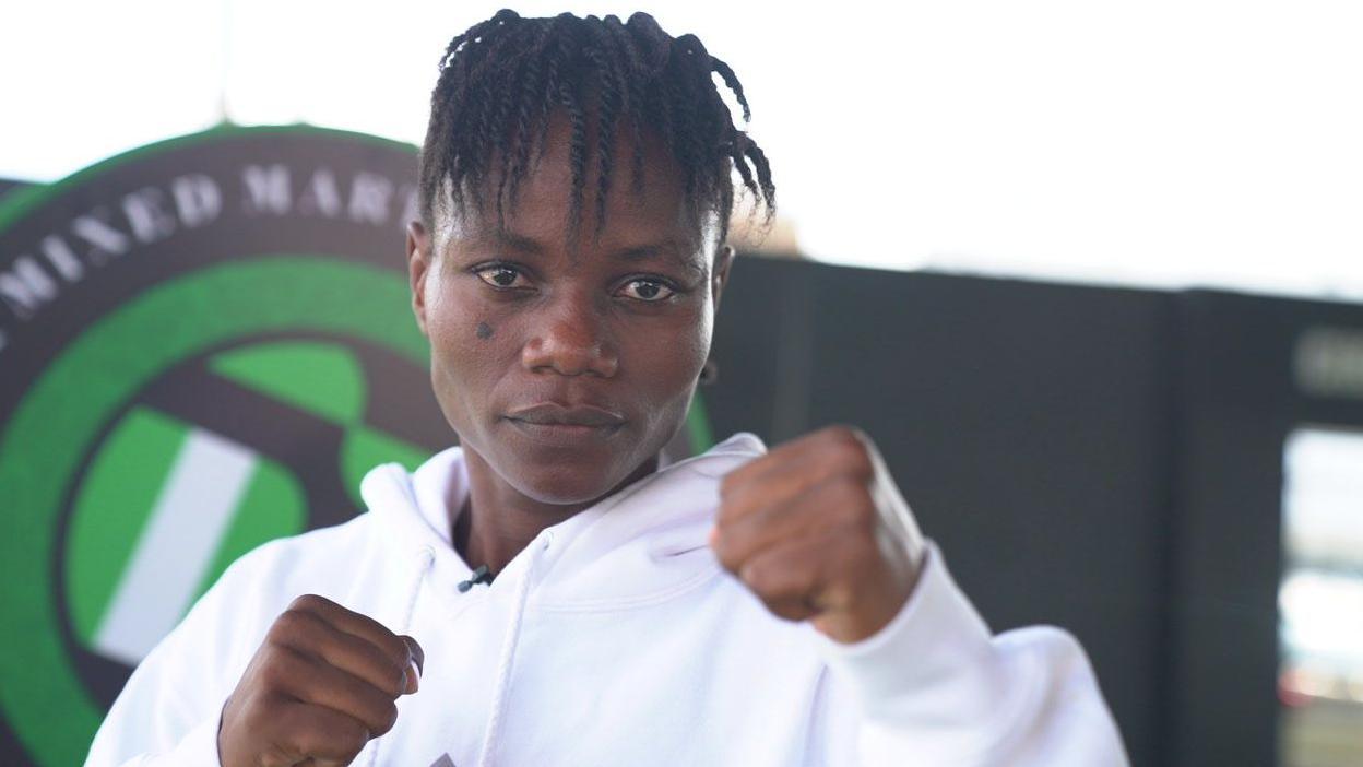 MMA fighter Juliet Ukah wears a white top and holds up two fists as she looks straight