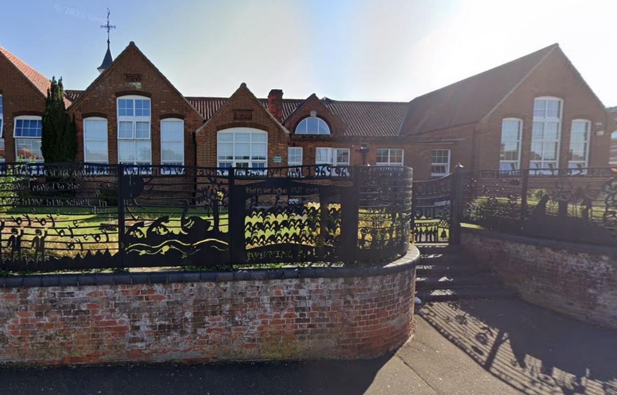 Black fences surround Fakenham Juniors, with grass in the foreground of the red brick building.