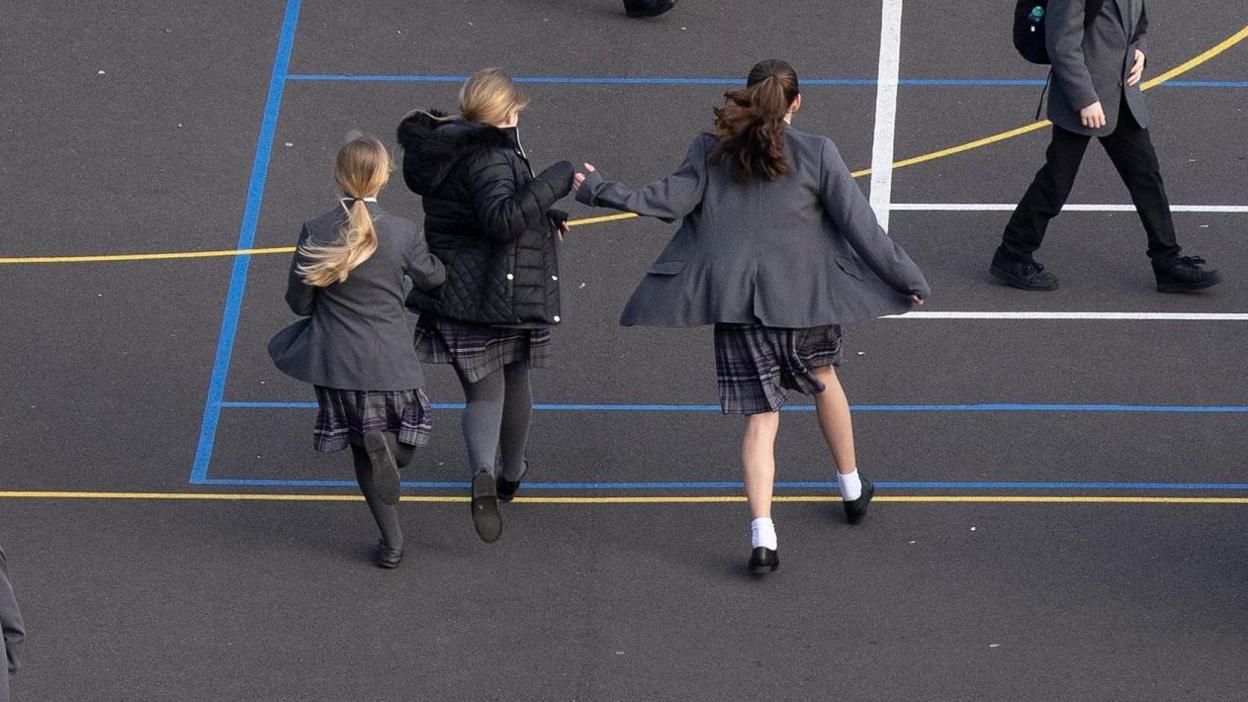Children playing in a Surrey secondary school. Three girls run across a playground. In the background a boy walks past.