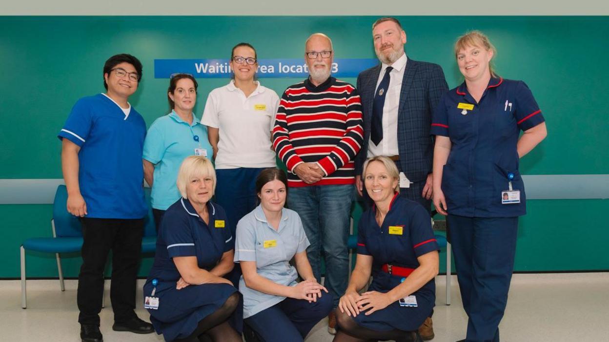 A group of men and women in hospital clothing around a man with grey hair and a beard in a striped blue and red top with his hands clasped in front of him