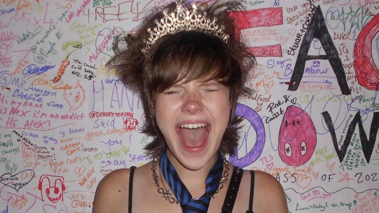 A girl with a pixie cut and brown hair wearing a tiara. She appears to be screaming with her eyes closed and also wears a necktie and silver necklace.
The background is full of colourful scribbles and messages. 