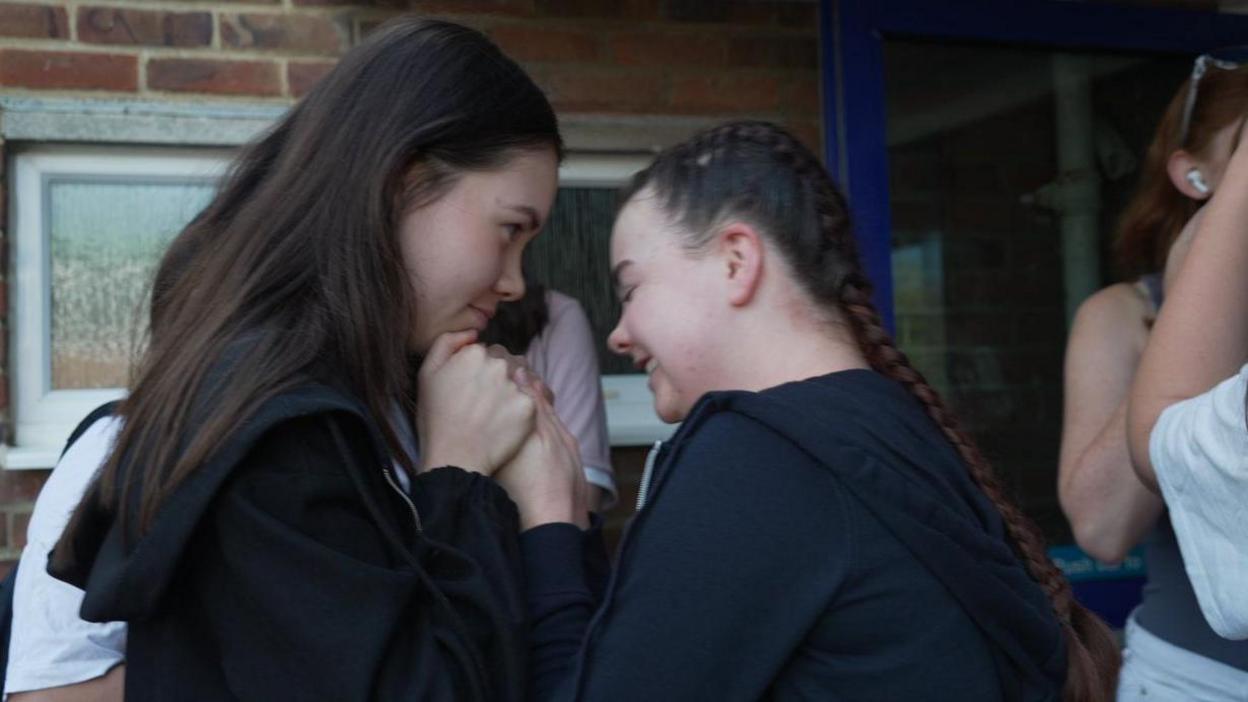 Two female friends hold hands, looking at each other. They appear to be nervously waiting outside their college. The young woman on the right is smiling and looking down, she has her dark brown hair tied back in long french plait braids and wears an unzippled black hoodie. Her friend on the left is holding her hands and looking at her. She has long brown hair and also wears an unzippled black hoodie.