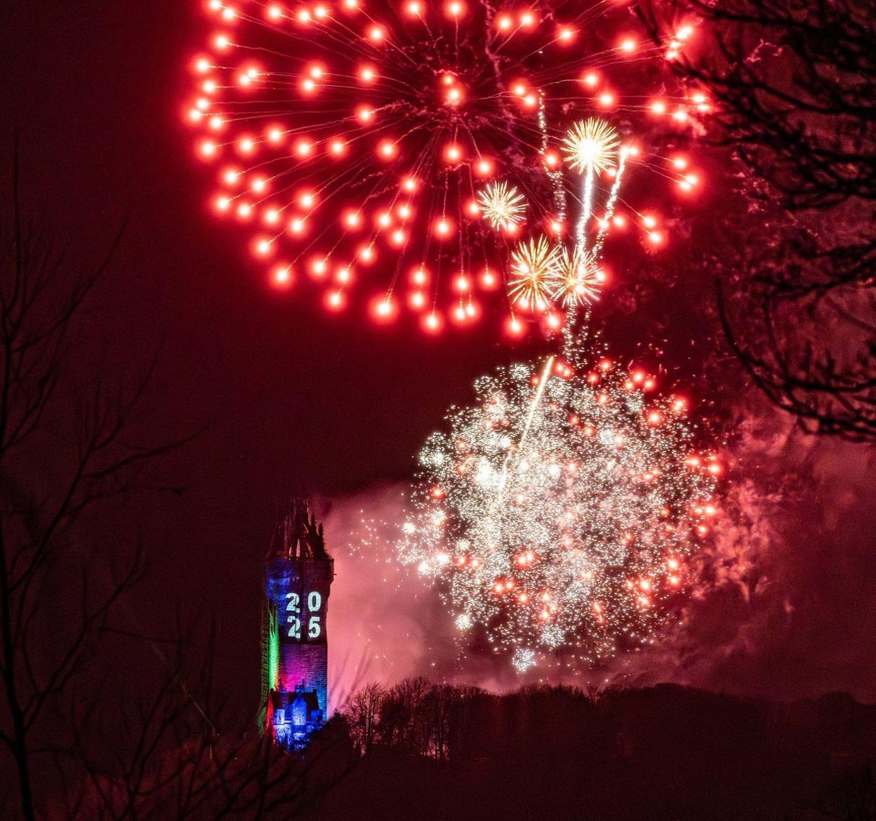 Red and white fireworks explode near the Wallace Monument, a tall old tower with 2025 projected on it.