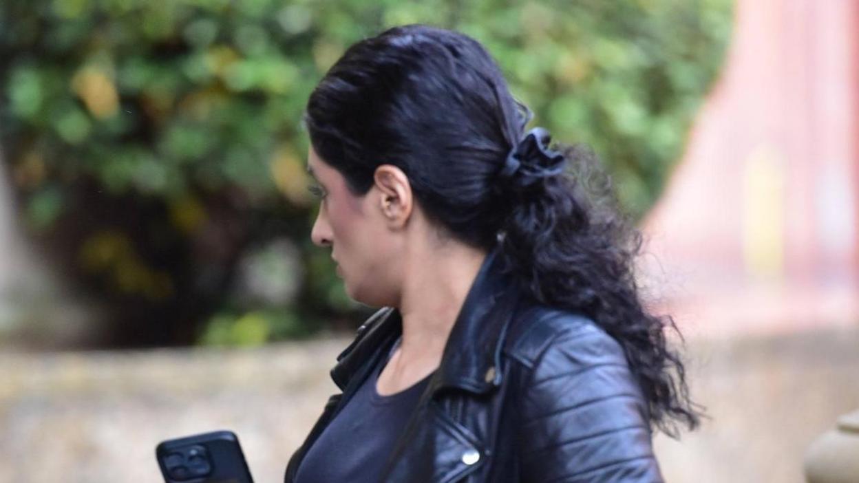 A woman with long dark hair and wearing a dark leather jacket walking outside court - she has her head turned away from the camera and a phone in her hands