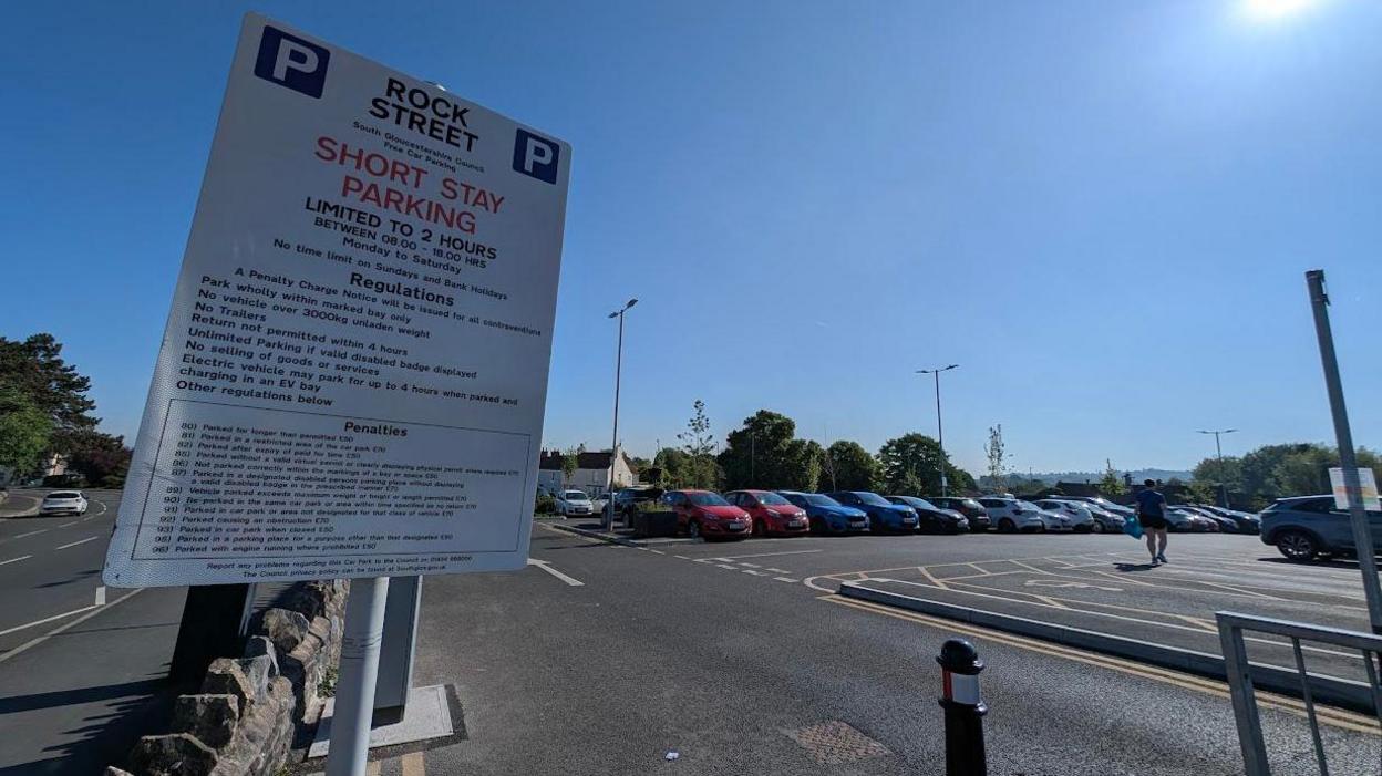 Cars parked in a car park. The sky is blue. The cars are on the right, on the left is a wall saying Rock Street short stay parking.