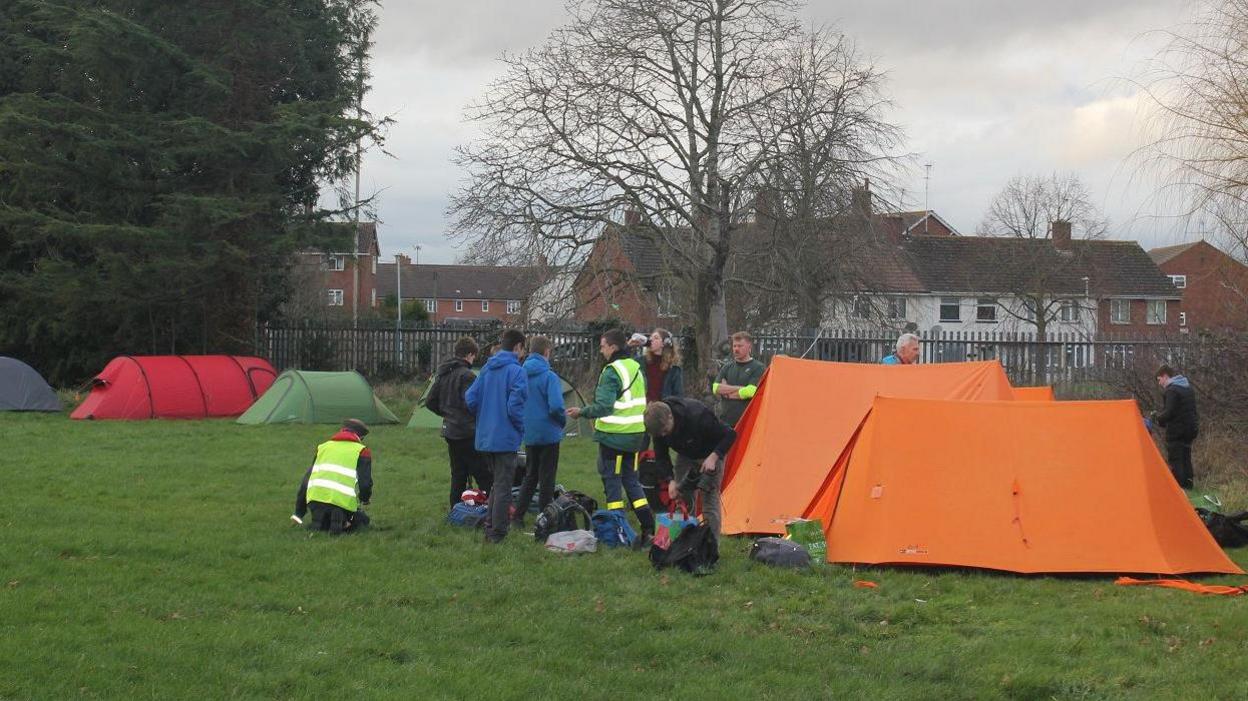 A group of people, some of whom are wearing reflective gear, gathered around tents on a field in daylight.