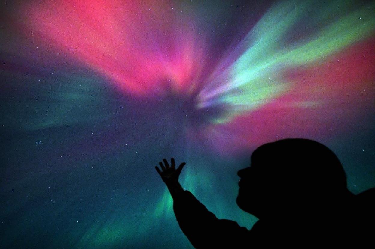 Silhouette of a person reaching to the sky with one hand. The sky is lit with different colours including shades of red, purple, green and blue.