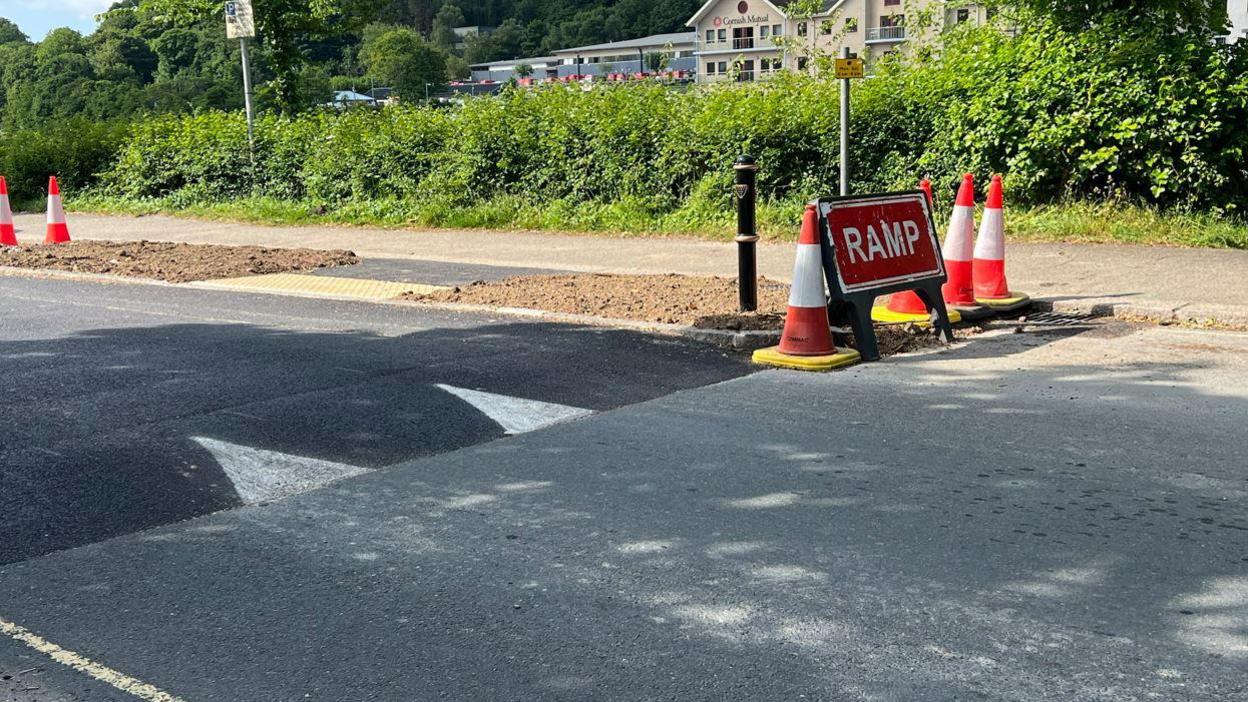 A table-top speed hump which forms part of a crossing installed on Malpas Road in Truro. Several traffic cones and a red warning sign saying ramp are on one side of the road. Buildings can be seen in the background past a hedgerow.