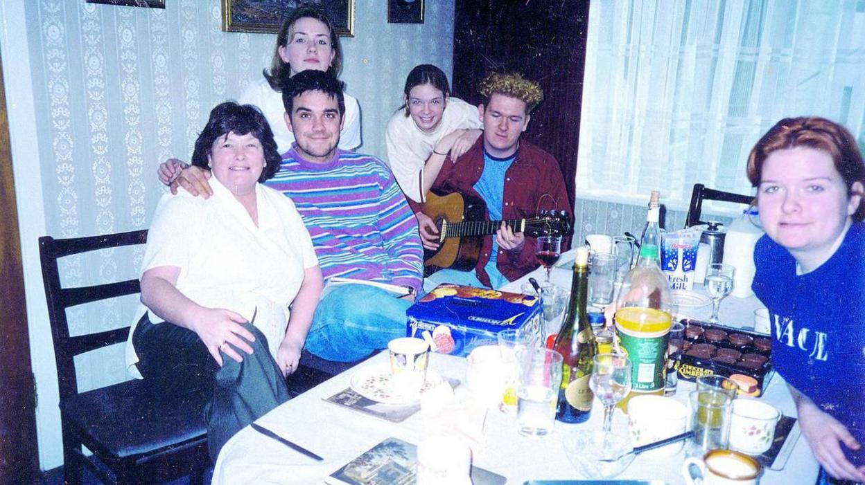 Robbie Williams in a stripy jumper next to a man with curly hair playing the guitar. they are sitting around a table on which biscuits, cups of tea, a wine bottle and several wine glasses are visible. Also around the table are four women