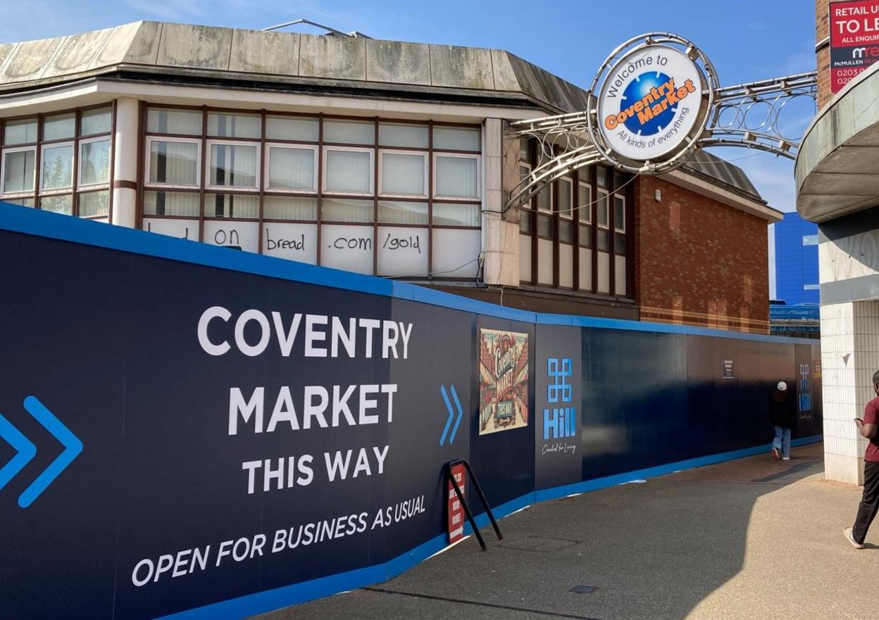 A blue and white hoarding sign including the words Coventry Market this way is in front of a building.
