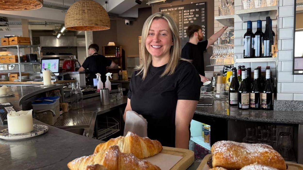 Llun o Nia Roberts wrth gownter ei bwyty - Baravin - gyda croissants ar y cownter ac aelodau staff yn parhau a'u gwaith yn y cefndir