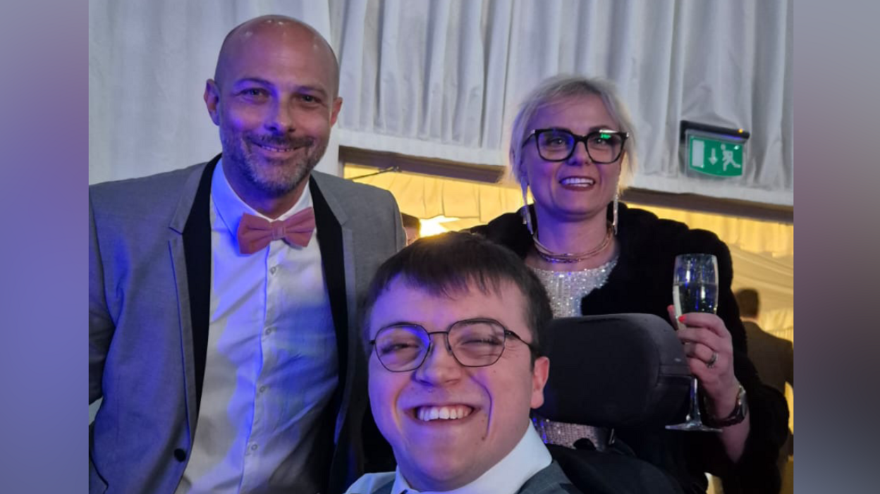 Mike and Nikki Kemp smile at the camera while standing above Harrison. Mike he bald and has a beard. He wears a grey suit with a white shirt and pink bow tie. Nikki has white blonde hair that is tied back, glasses and a silver sequin dress with a black coat over the top. 