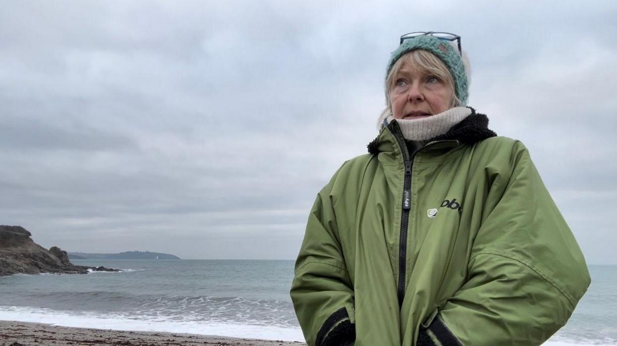 Mrs Hitchcock standing on the beach with the sea is behind her. She is wearing a green winter jacket, a beige scarf, and a blue woolly hat. 