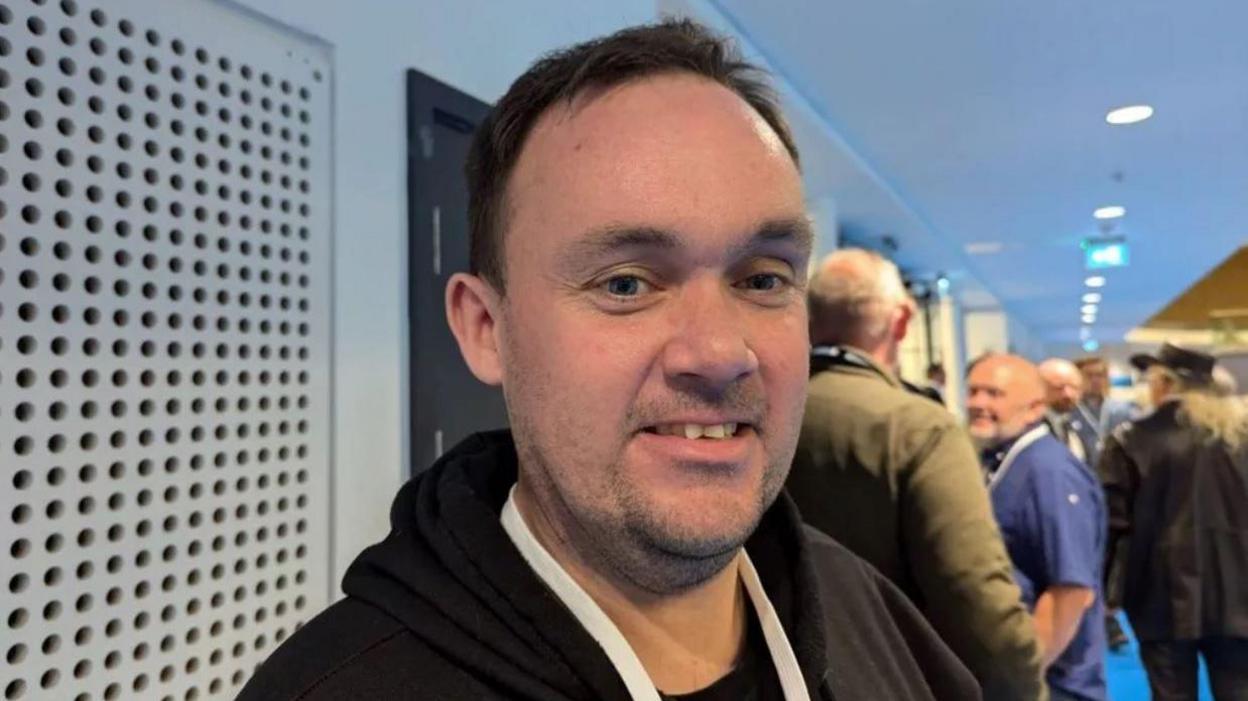 Image of Jonathan. He is wearing a black hooded jumper and is stood in front a number of people.