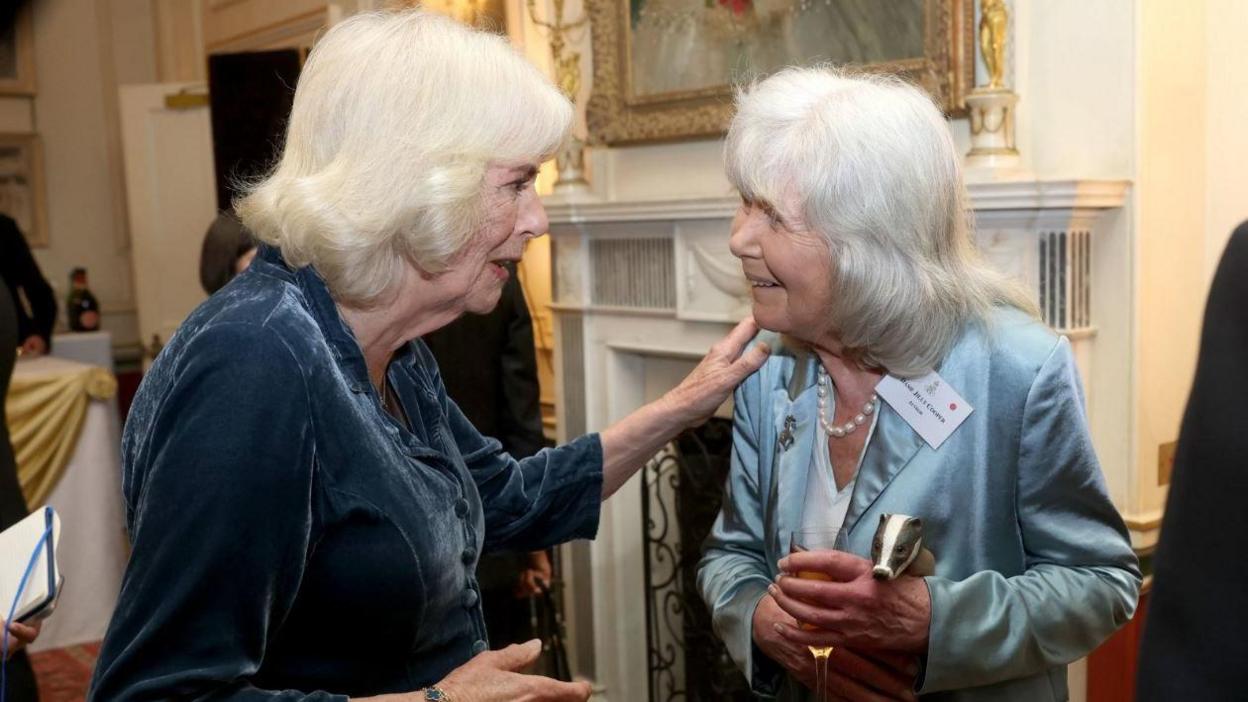 Queen Camilla and Dame Jilly Cooper at a reception at Clarence House. Dame Jilly wears a pale blue silk suit, while the Queen wears a dark blue velvet jacket. A grand room with lots of other people is visible in the background. 