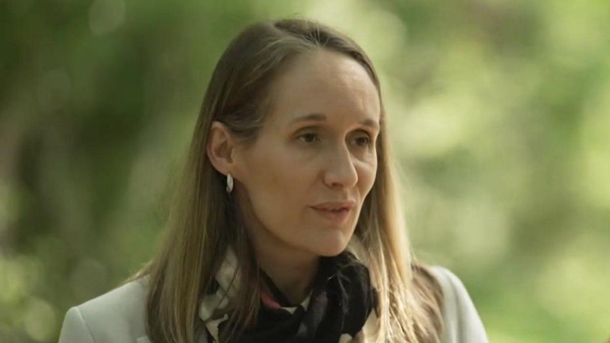 A woman with long fair hair is standing in a forested area. She is wearing a light coloured jacket with a cream and black scarf. 