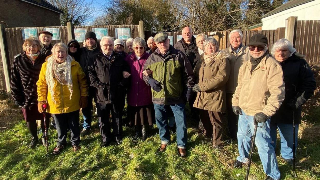 Residents who live on Titford Road in Oldbury. There are around 17 people standing together. Mostly older people. They are standing in front of protest signs on a grassy embankment. It is winter time so they have coats on.