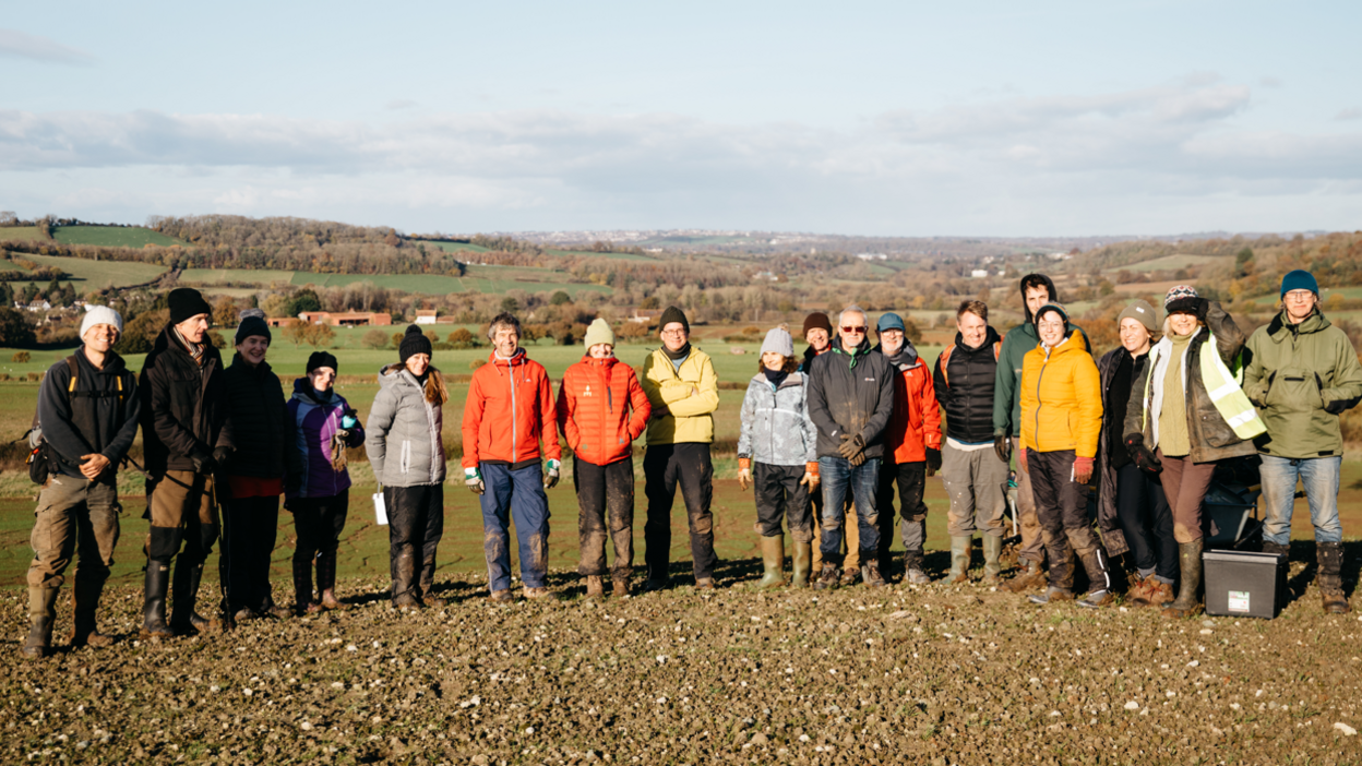 A group of people in outdoor gear stand in a field on a cold but sunny day. Small patches of woodland can be seen in the background.