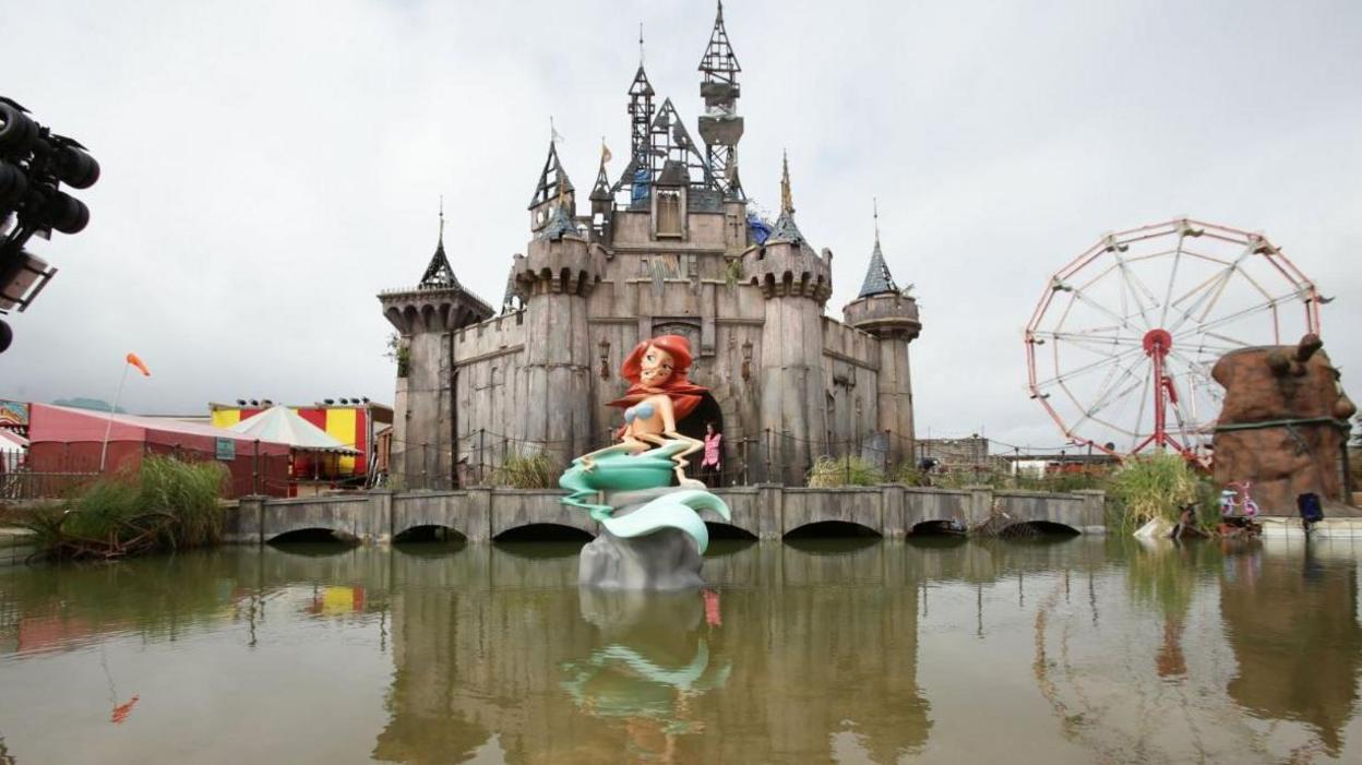 A sculpture of a derelict version of the Disney castle, with a distorted model of the Little Mermaid sitting on a lake in front. A big dipper and circus tents are visible in the background.