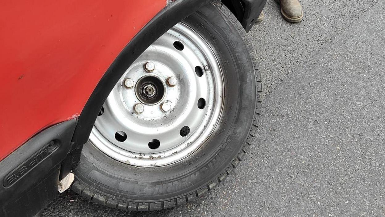 This image was taken after the horsebox broke down on the road. The focus is on a detached wheel that has come off the vehicle. The wheel has a silver steel rim with circular holes and a black tire. It’s lying on a road and the horsebox’s red body and black fender are visible, indicating where the wheel came from.