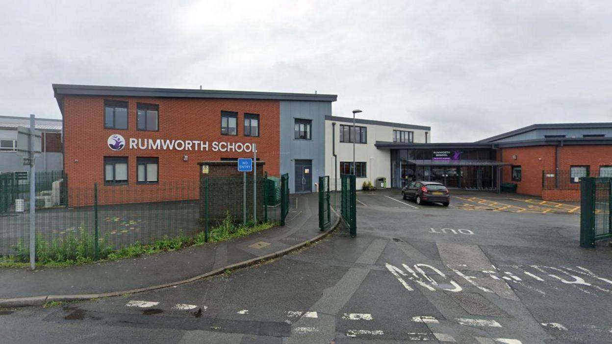 View from the road of Rumworth School. It is a modern red-brick building with grey and purple signage. Reception is visible from afar. The car park gates are open and there is one parked car. 