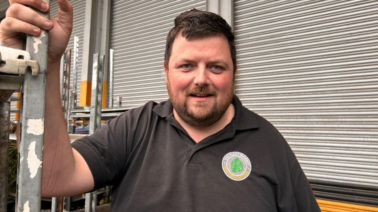 Plant Market owner Sean Thomas, who has short brown hair and a beard, is wearing a black T-shirt and stands in front of closed roller shutters in a warehouse