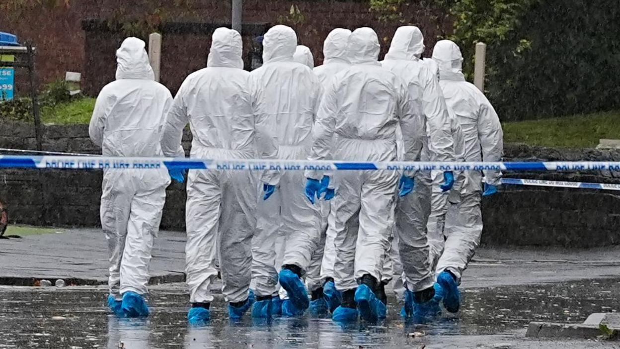 A group of forensic investigators in white PPE and blue gloves and shoe coverings, walk away from the camera behind a line of blue and white police tape. 