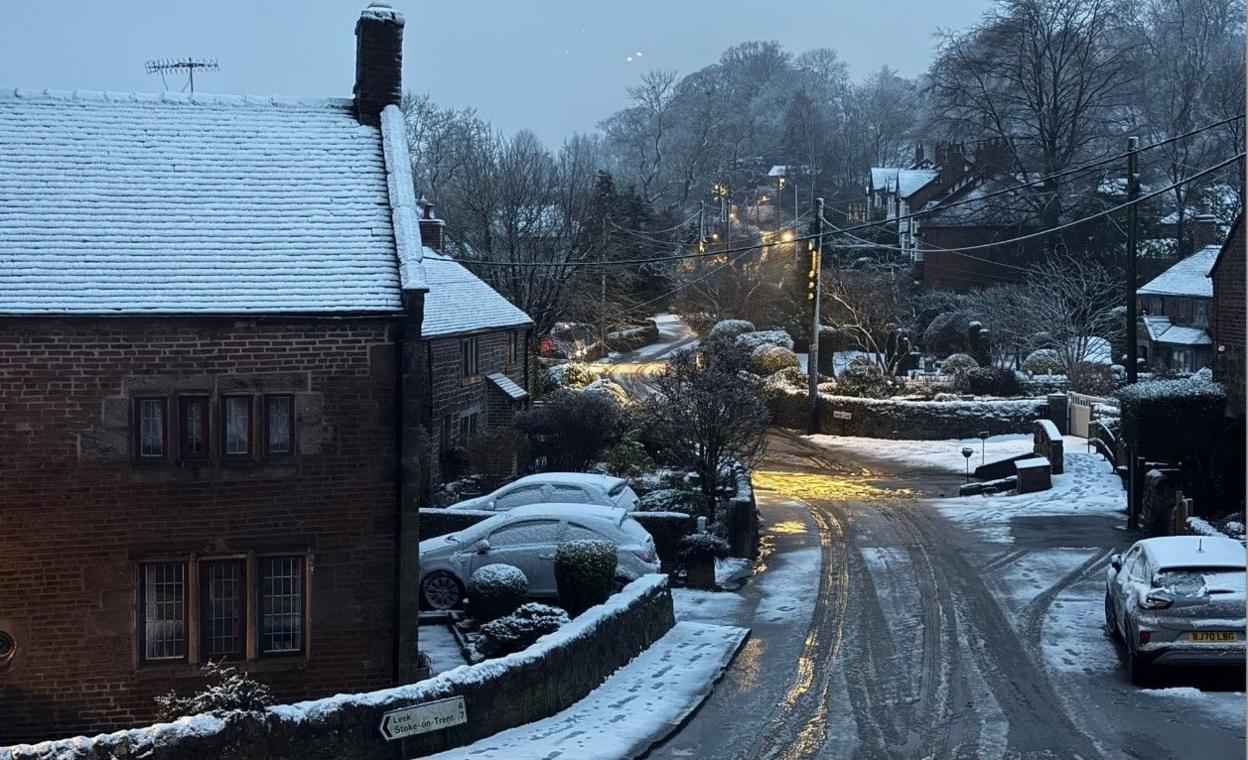 A road on the right of the photo. The pavement to the left of the road on the image has snow on it, as does the roof on the building behind it on the bend. Parked cars have snow on them. 