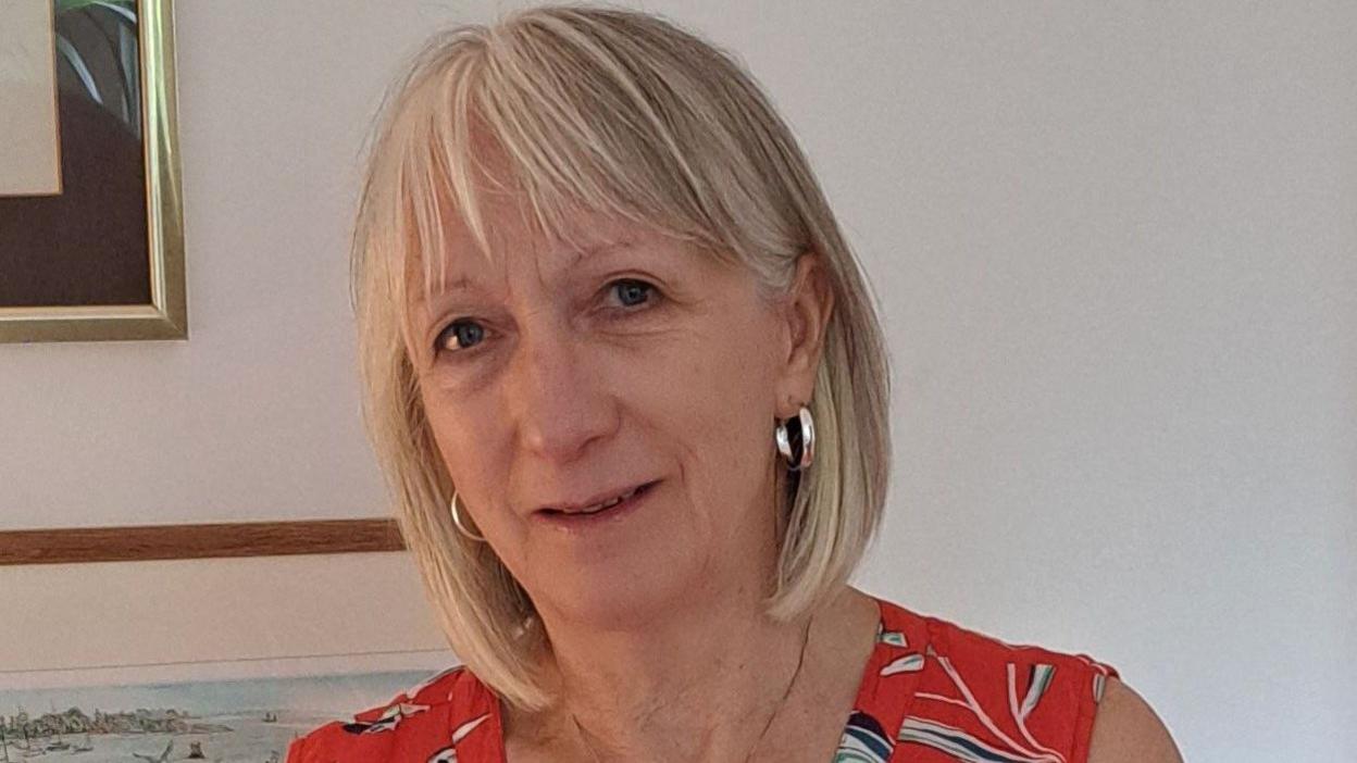 A woman with blonde hair is smiling at the camera and is wearing a red patterned top. 