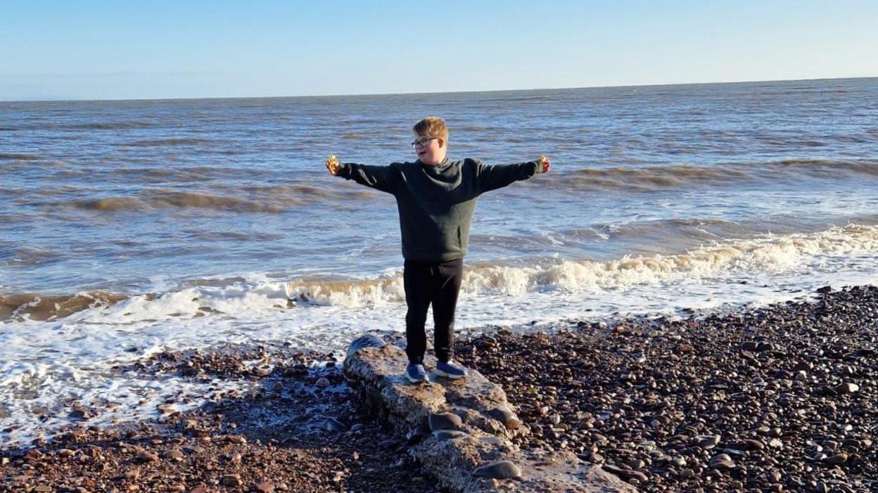 A boy is standing in front of the sea with his arms outstretched.