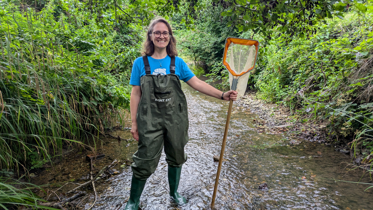 Ms Brookes is standing in the stream with a large fishing net on the end of a wooden pole - about the same height as her. She is wearing green wellies and a green waterproof overall. She has long hair, glasses and is smiling.