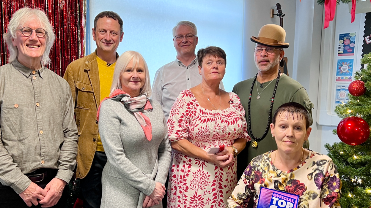 A man with long grey hair, grey shirt and glasses, a man with brown hair and a yellow top, a woman with grey hair and a long grey dress, a man with grey hair and a white shirt, a woman with dark hair and a red and white patterned dress, a man with a green tshirt and brown bowler hat and a woman with short dark hair and a floral patterned dress, holding a blue and pink trophy