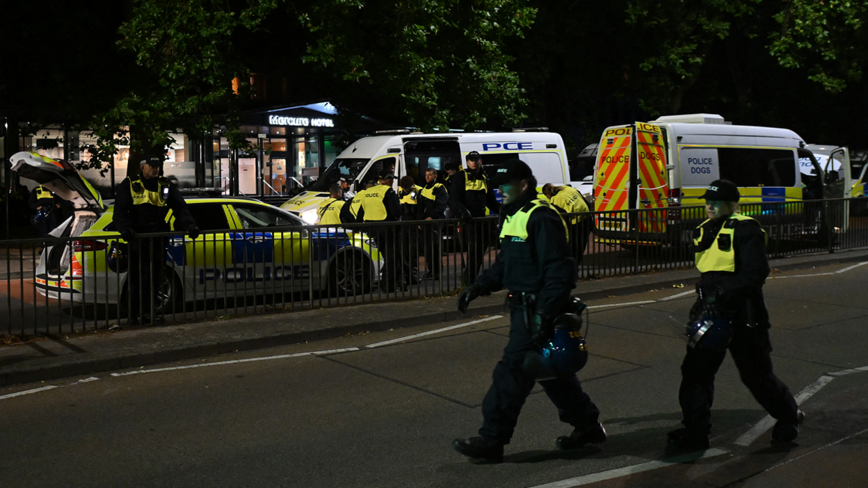 Two police officers wearing high-vis jackets walking across a road, with police cars, vans and further officers in the background.