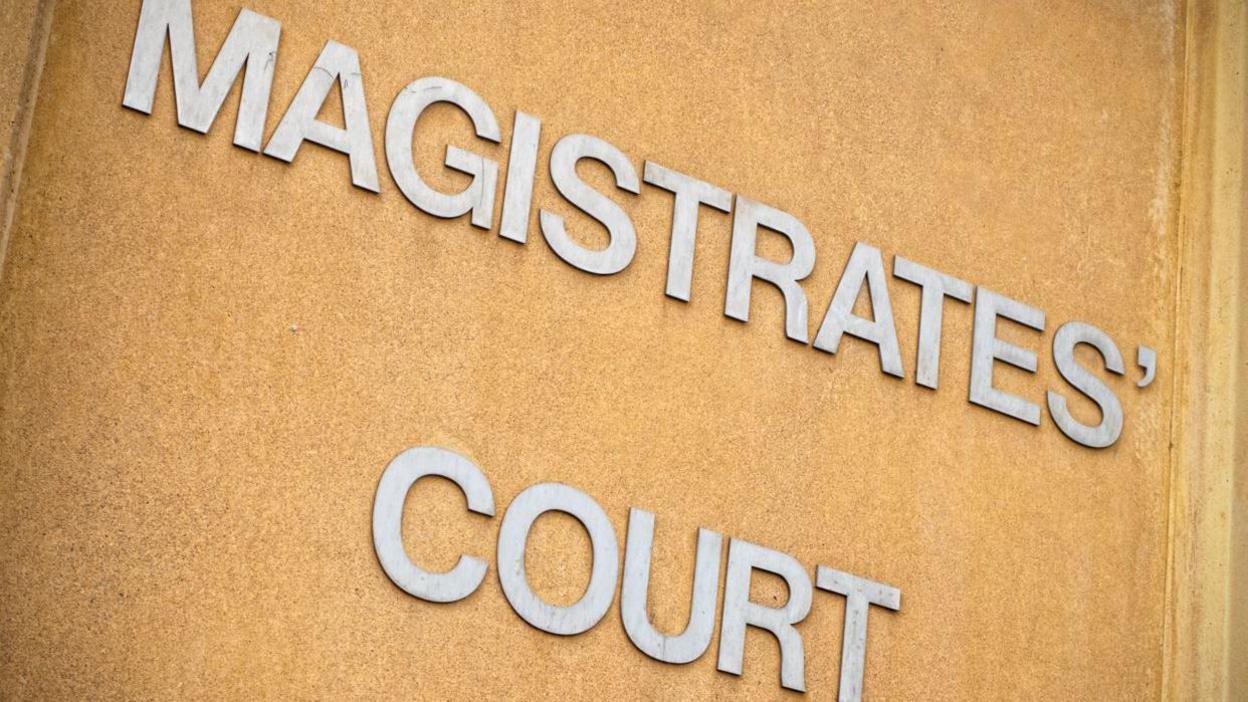 A large shiny metal sign on a brown wall which reads Magistrates Court